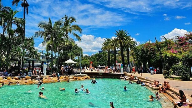 A pool area with people swimming and sunbathing, surrounded by palm trees and a clear sky. - 2bedrm unit next to southbank (Highgate Hill)
