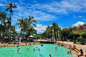 A pool area with people swimming and sunbathing, surrounded by palm trees and a clear sky.