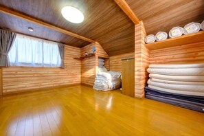 A log cabin room with a wooden floor, a window with curtains, a bed with pillows, and a shelf with folded blankets. - SOMEDAY (Onjuku)