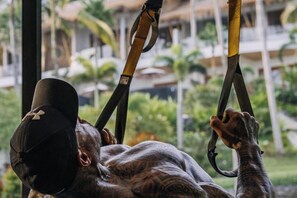 A person hanging from gymnastic rings in a room with a view of palm trees and a building in the background. - Neano Escape (Bali)