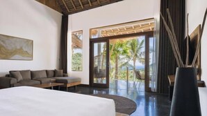 Habitación de hotel moderna con una cama grande, un sofá y vistas a palmeras y al mar a través de grandes ventanales.