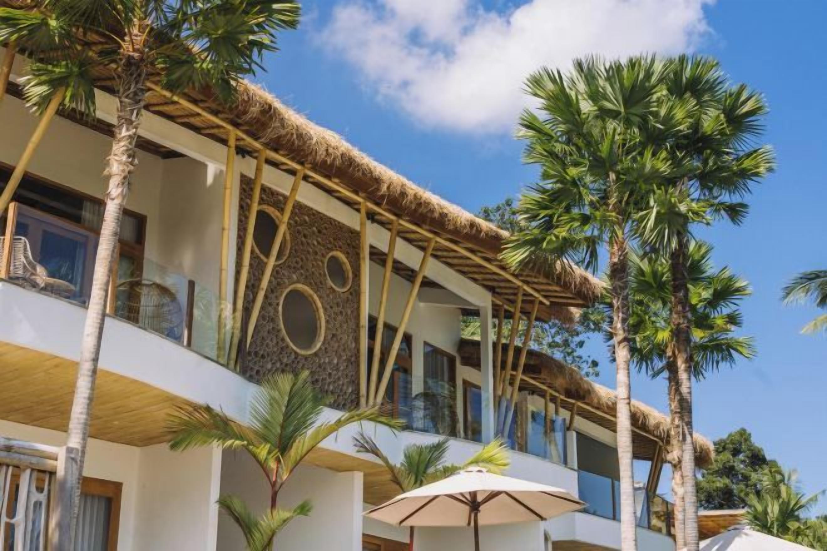 A multi-story hotel with a thatched roof, palm trees, and outdoor umbrellas.