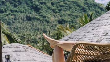 A wicker chair with a person sitting, overlooking a lush green landscape and a thatched roof structure.