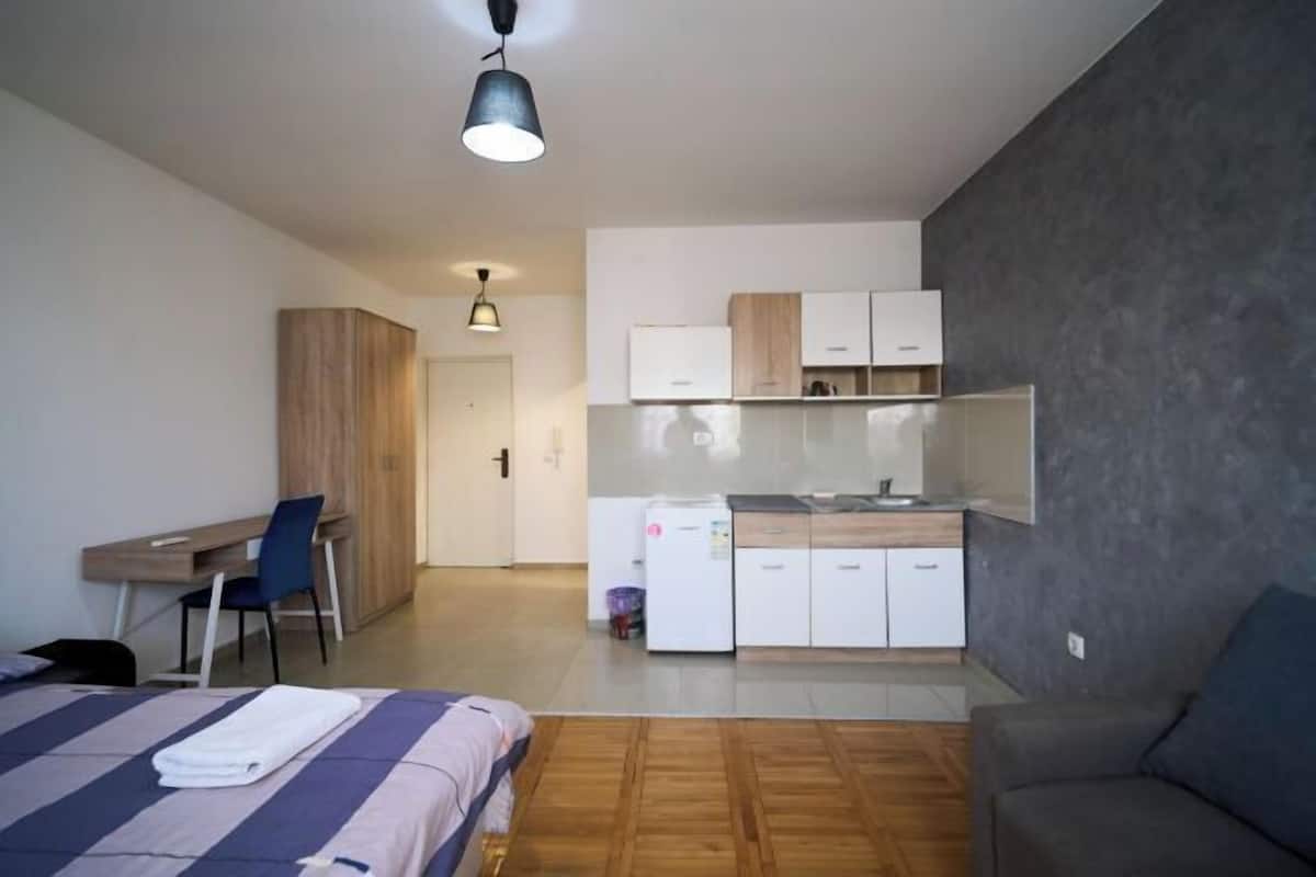 A modern bedroom with a bed, a desk, a chair, a wardrobe, and a kitchenette.