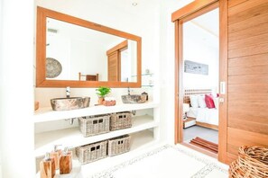 A bathroom with a large mirror, a sink, wicker baskets, and a bed visible through an open door. - Villa ARNAUD 17 (Bali)