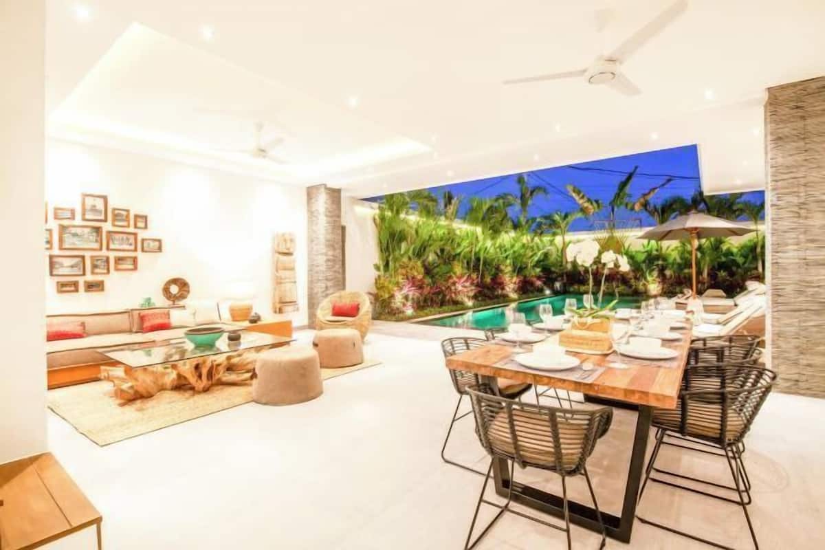 A modern living room with a pool view, a dining area with chairs and a table set for two, and a ceiling fan.