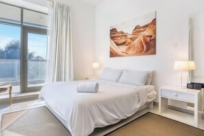 A hotel room with a bed, a nightstand, a lamp, a painting, and a view through a large window. - bnbme Great Value Apt in Dubai Sports City (Dubai)