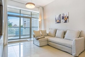 A modern living room with a sectional sofa, a large window, and a wall-mounted lamp. - bnbme Great Value Apt in Dubai Sports City (Dubai)