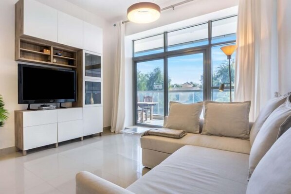 A modern living room with a large window, a flat-screen TV, and a sofa. - bnbme Great Value Apt in Dubai Sports City (Dubai)