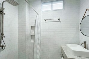 Ein modernes Badezimmer mit weißen, rechteckigen Fliesen, einer Dusche mit Handbrause, einer Duschkabine aus Glas, einem weißen Waschbecken inklusive Spiegel und einem Handtuchhalter.