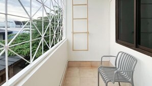 A balcony with a view of greenery, a window with a metal grid, a wooden ladder, and a gray chair.