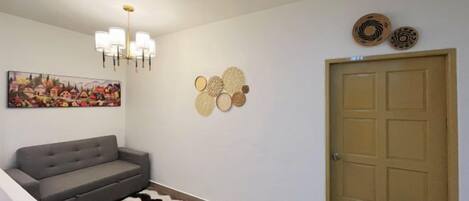 A hallway with a grey sofa, a patterned rug, a chandelier, wall art, and a door.