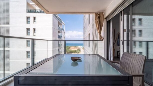 A balcony with a glass table, a chair, and a view of buildings and the sea. - Achziv Beach Haven 3BR Apartment by Sea N' Rent (Nahariyya)