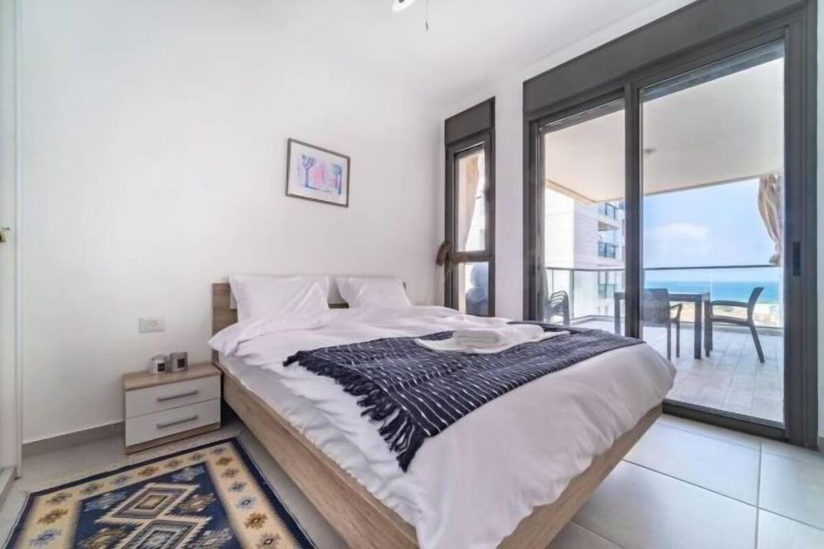 Un dormitorio con cama, mesita de noche, alfombra y balcón con vistas al mar.