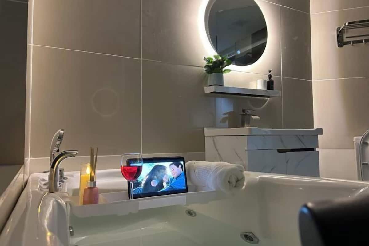 Een moderne badkamer met een bad, een ronde spiegel, een klein plankje met een plant en een tablet waarop een afbeelding staat.