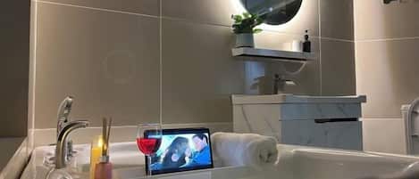 Uma casa de banho moderna com uma banheira, um espelho redondo, uma pequena prateleira com uma planta e um tablet mostrando uma imagem.