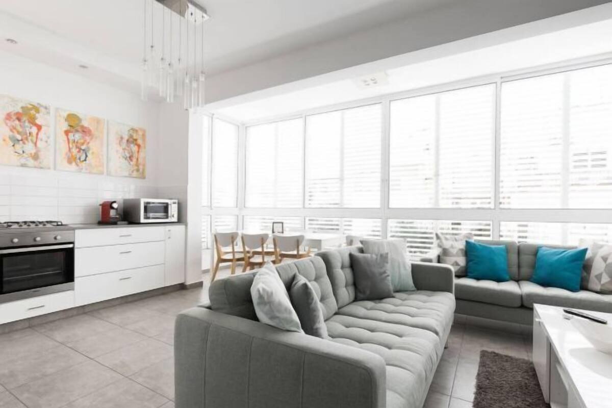 A modern kitchen with white cabinets, a gray sofa, and a dining area with chairs and a table.