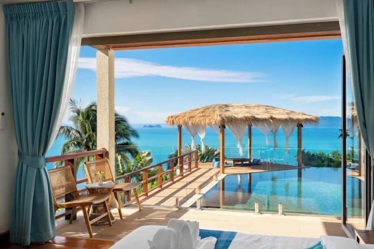 A room with a large glass window overlooking a pool and the sea. There are wooden chairs and a bed with white linens.
