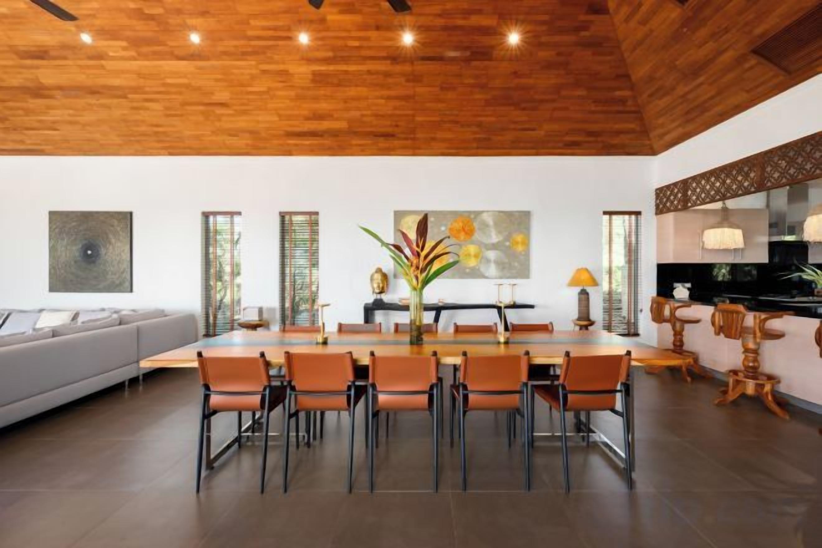 Un espace de repas moderne avec un plafond en bois, une table centrale entourée de chaises et un canapé à l’arrière-plan.