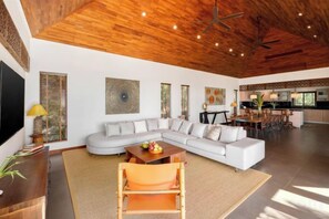 A spacious living room with a large sectional sofa, a wooden ceiling, and a kitchen in the background. - Baan Faasai 5-Bedroom Hillside Seaview Pool Villa (Koh Samui)