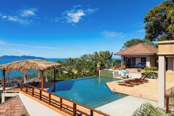 A pool area with a view of mountains, a thatched roof structure, and a building with a tiled roof. - Baan Faasai 5-Bedroom Hillside Seaview Pool Villa (Koh Samui)