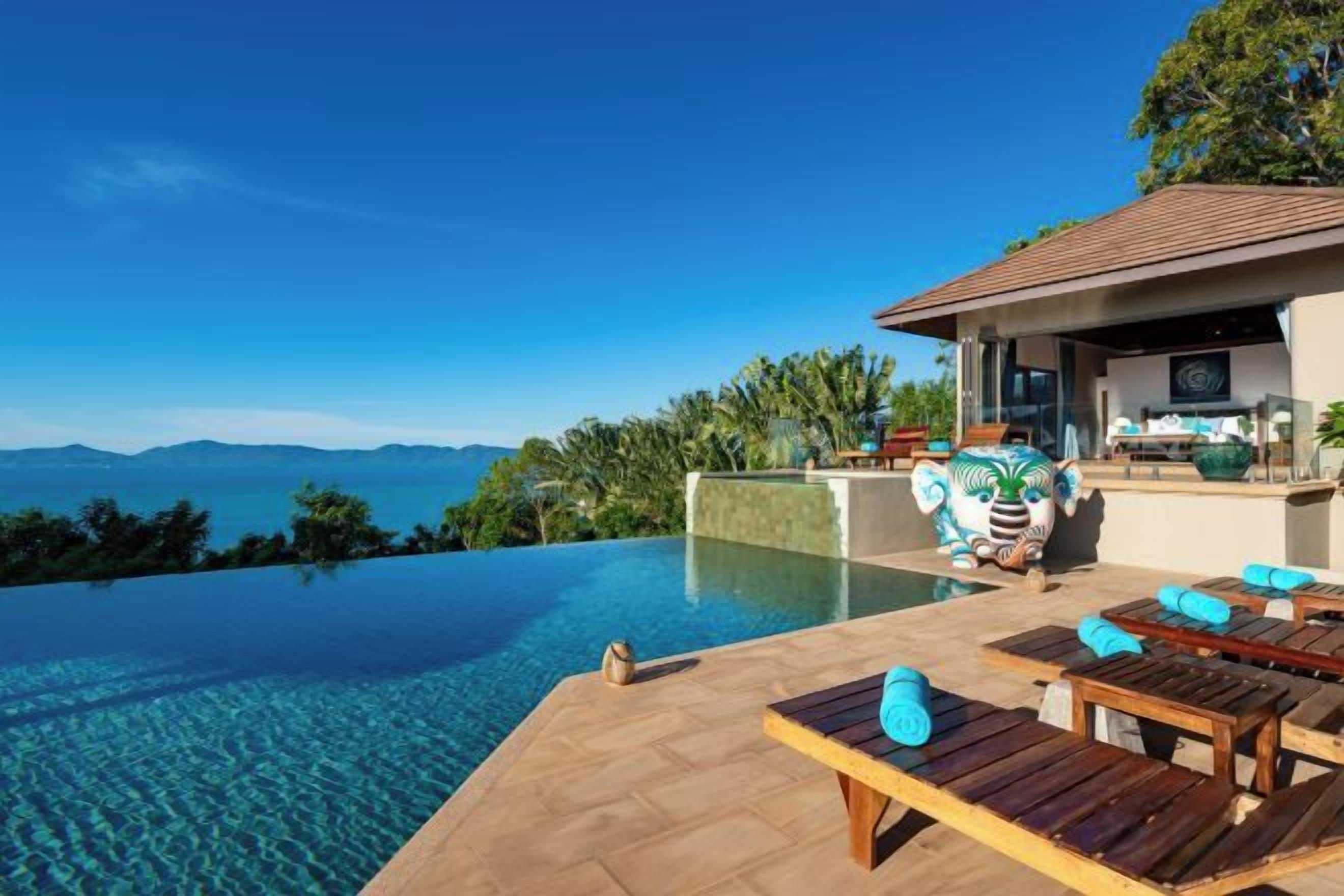 An infinity pool with a clear view of the mountains, wooden lounge chairs, and a covered seating area.