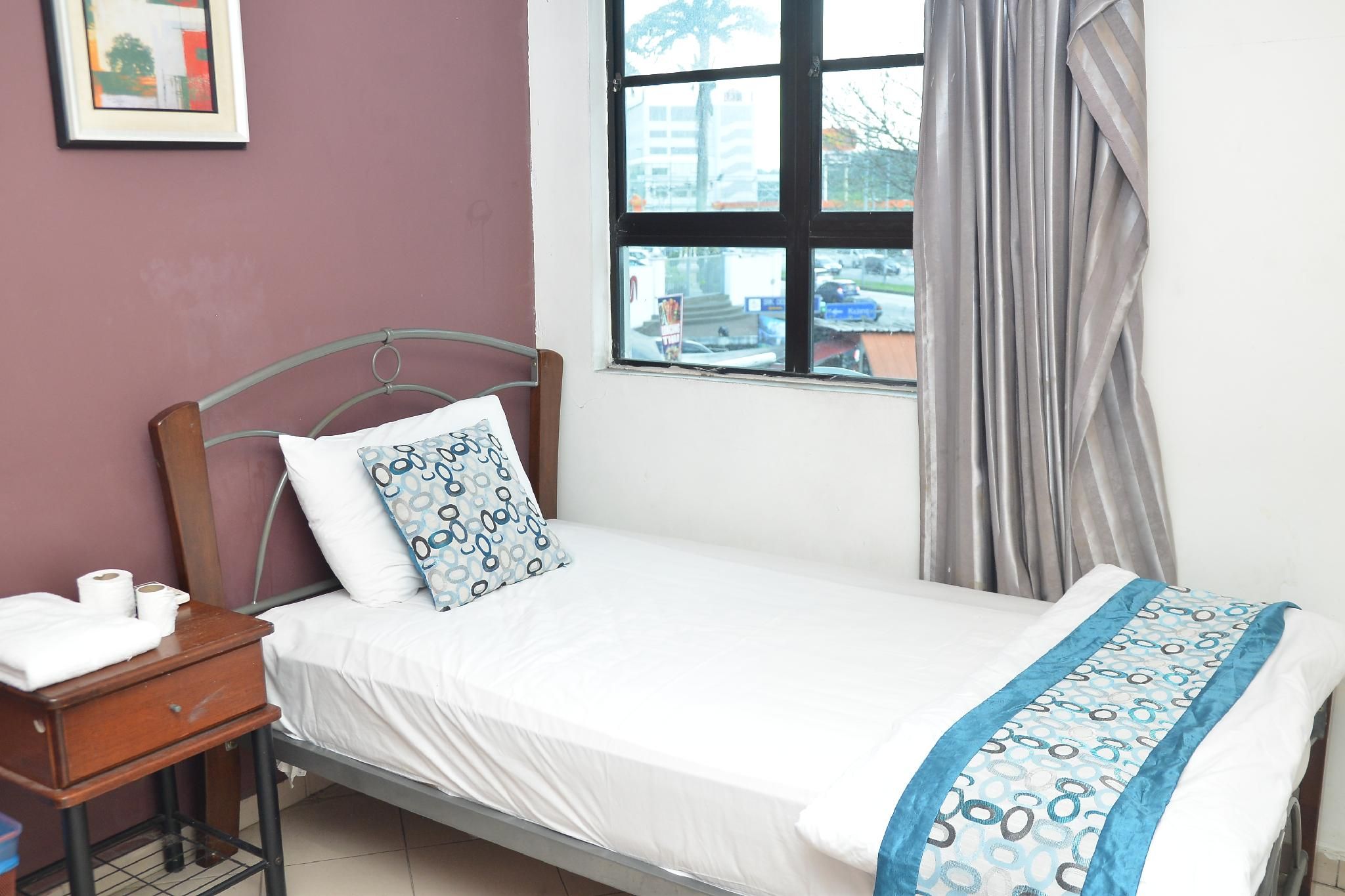 Uma cama de solteiro com colchão branco, almofada estampada, mesa de cabeceira com abajur e janela com cortinas.