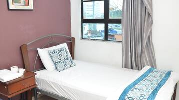 Uma cama de solteiro com colchão branco, almofada estampada, mesa de cabeceira com abajur e janela com cortinas.