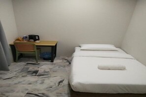 Een kleine kamer met een enkel bed, een bureau en stoel, een prullenbak en een betegelde vloer.