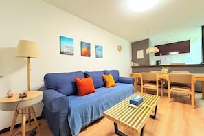 A modern living room with a blue sofa, a wooden coffee table, and a small round table with a lamp. There are three paintings on the wall and a kitchen area in the background.