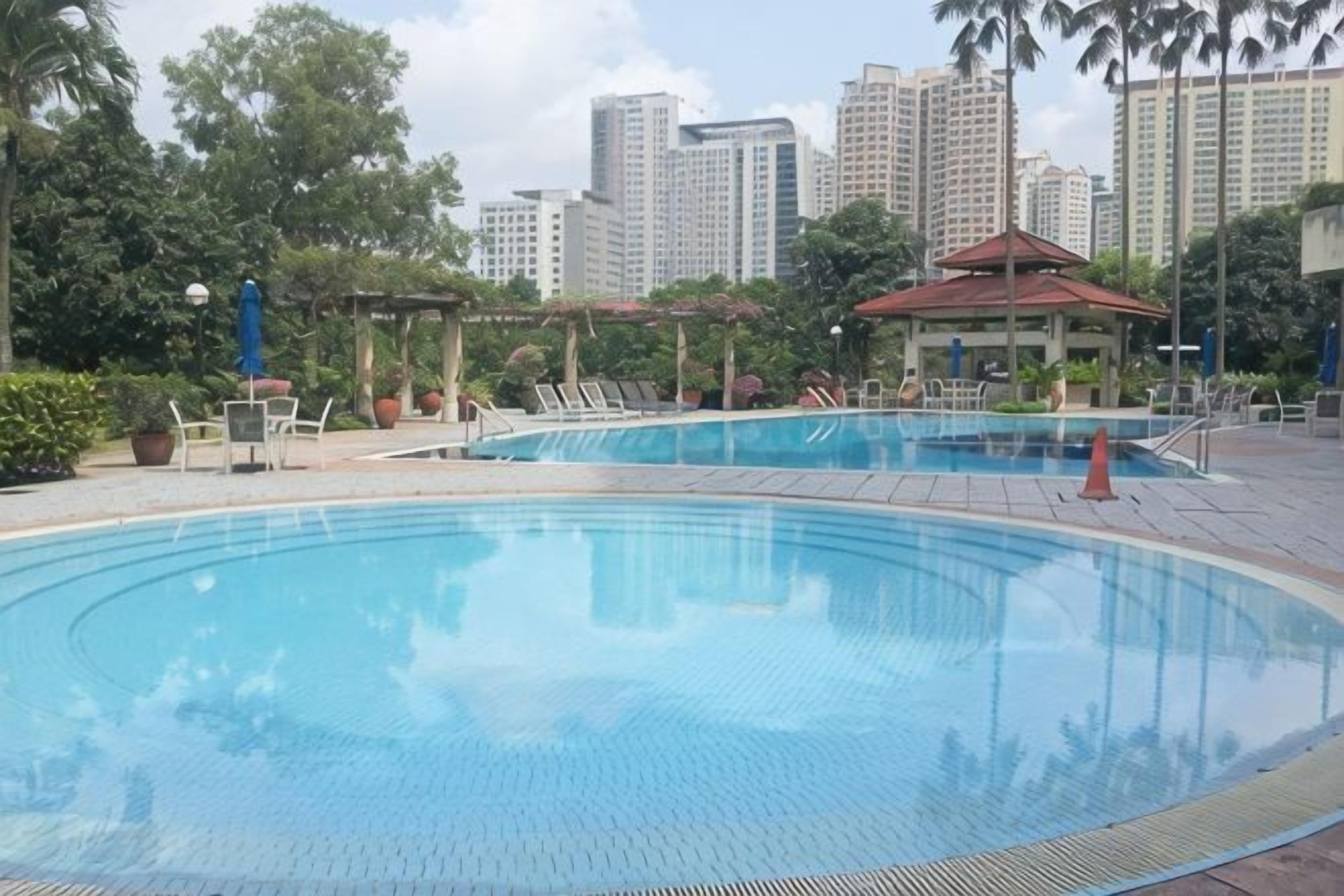 A swimming pool with lounge chairs and a gazebo, surrounded by greenery and tall buildings in the background.