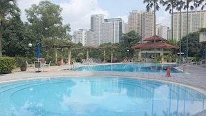 Una piscina con sillas de descanso y un pabellón, rodeada de follaje y edificios altos al fondo.