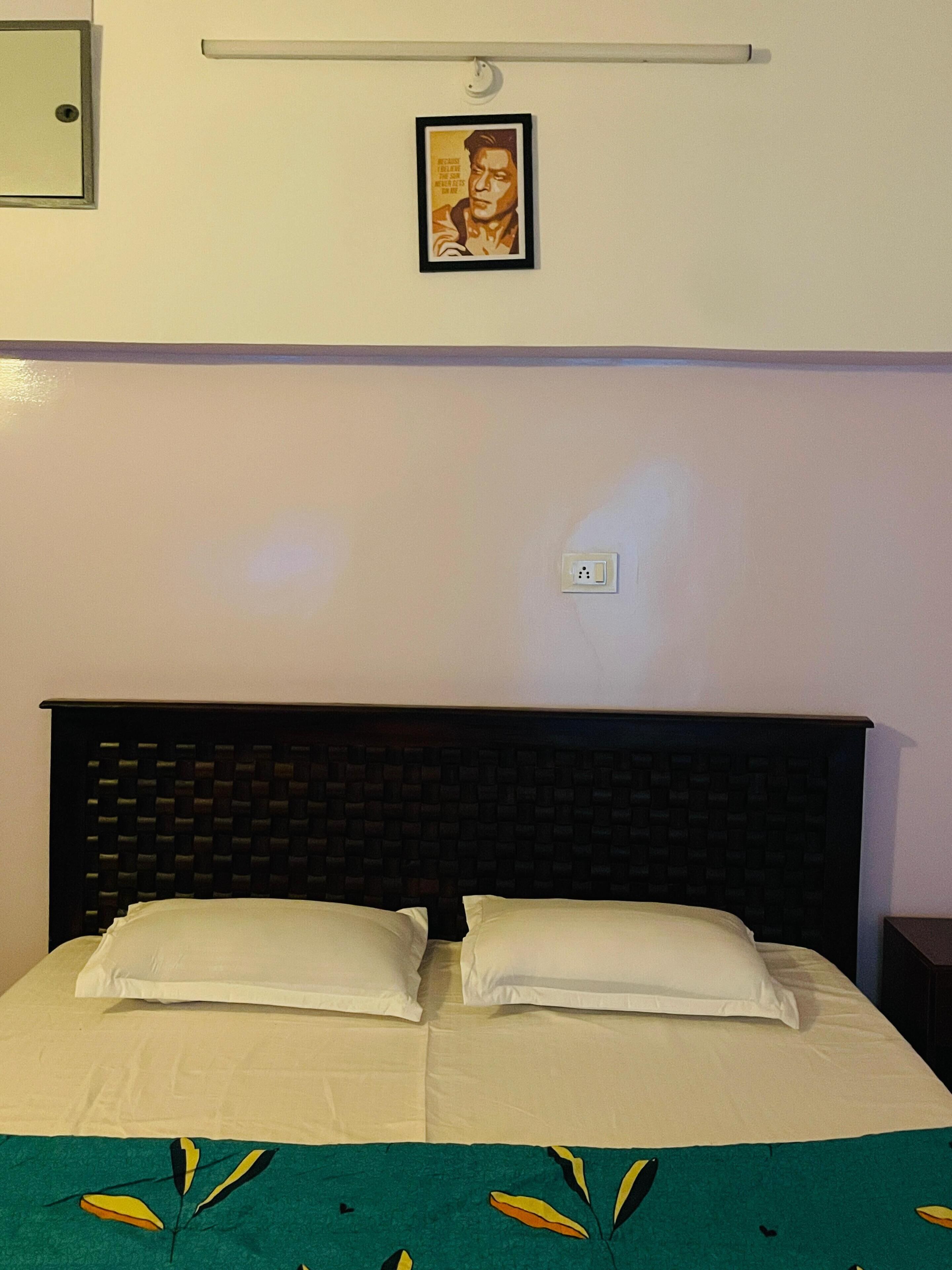 Ein Hotelzimmer mit einem Bett, zwei Kissen, einer grünen Bettdecke mit Blätermotiv, einem gerahmten Bild an der Wand und einer Steckdose.