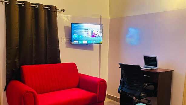 Une pièce avec un canapé rouge, un bureau avec un ordinateur portable, un téléviseur fixé au mur et un lit recouvert d’une couvre-lit à motifs.
