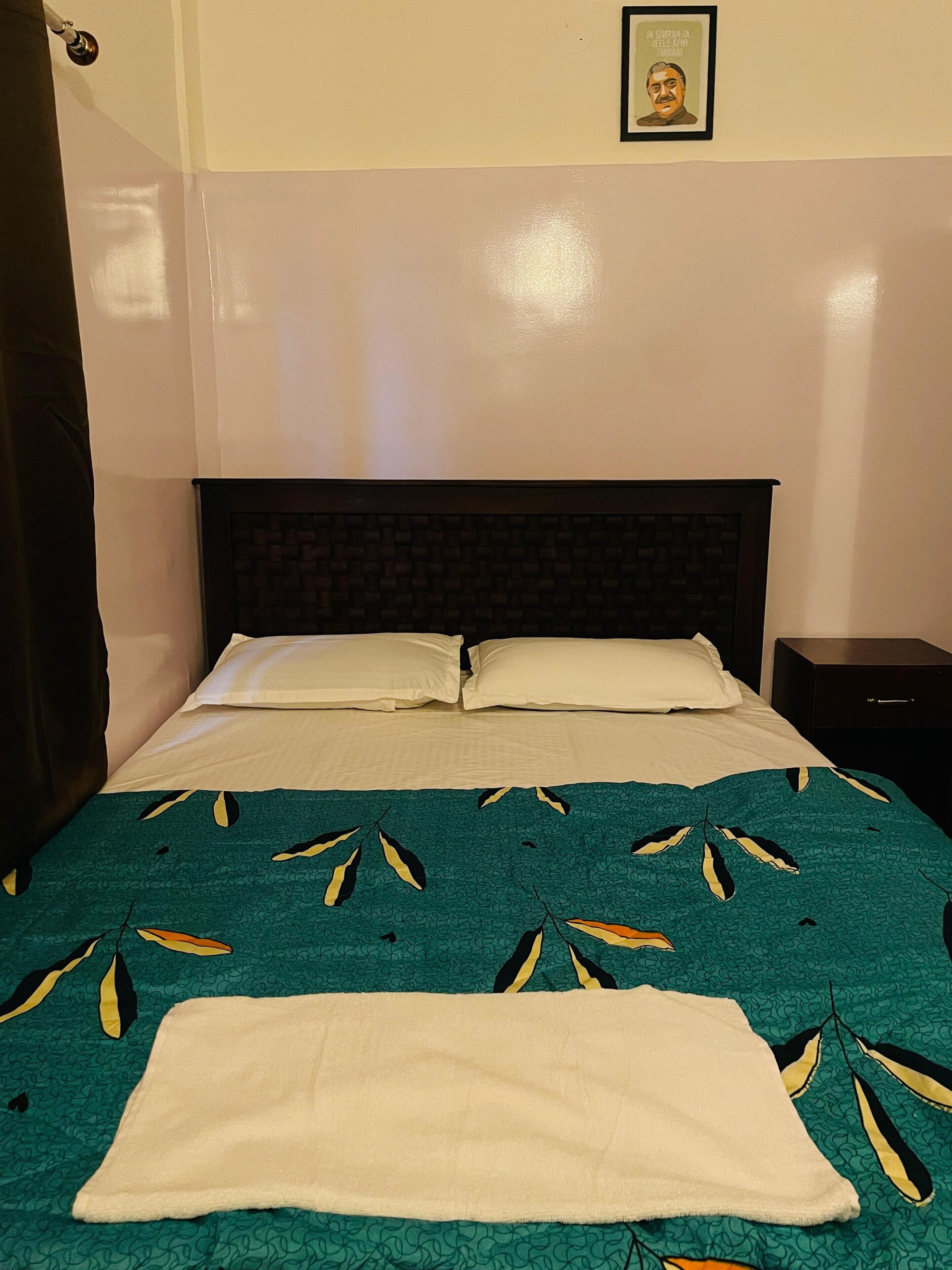 Un letto rifatto con cura, con una coperta verde acqua e gialla a motivi floreali, un cuscino bianco e un asciugamano bianco sul letto.