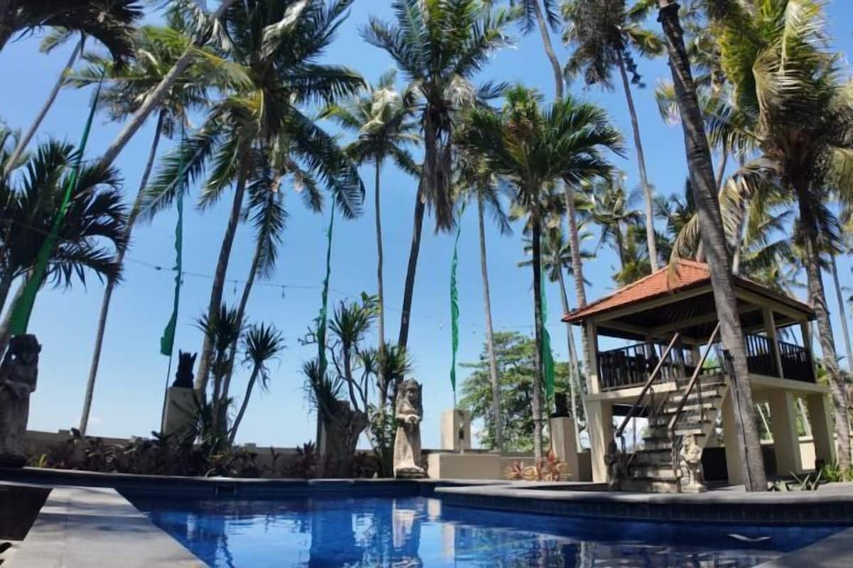 Un área de piscina con altas palmeras, una estatua de piedra y un pequeño pabellón.