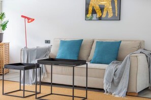 Una sala de estar con un sofá, dos mesitas auxiliares, una lámpara de pie roja y un cuadro enmarcado en la pared.