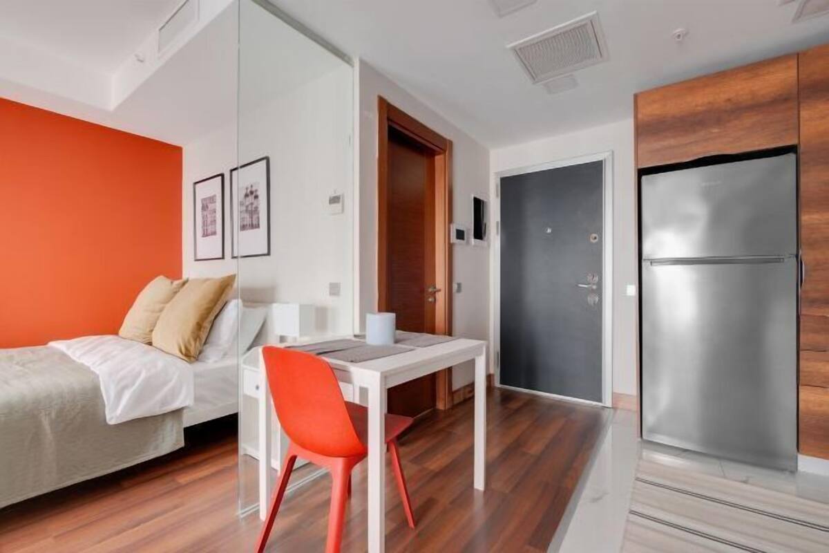 A modern room with a bed, a desk, a chair, a refrigerator, and a door.