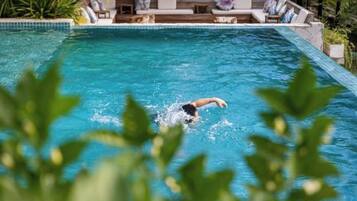 A swimming pool with a person swimming, surrounded by lounge chairs and a patio area.