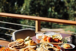 Una mesa con alimentos para el desayuno y un vaso de jugo, en un balcón con vista a árboles.