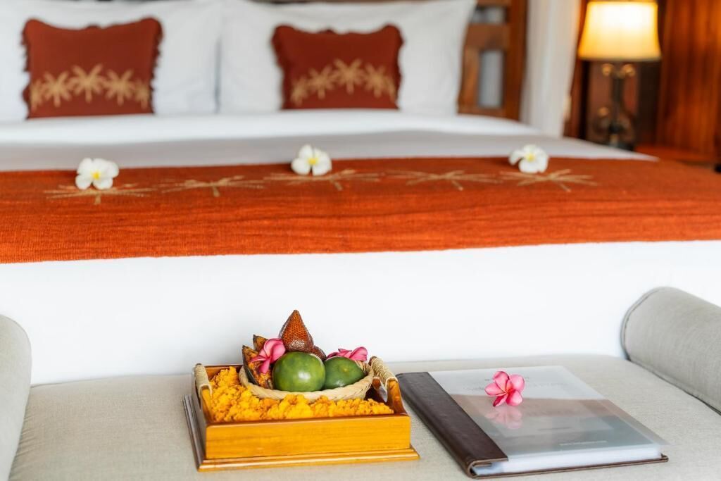 Una habitación de hotel con cama, una mesita de noche con un libro y una flor, y un cuenco decorativo con frutas y flores.