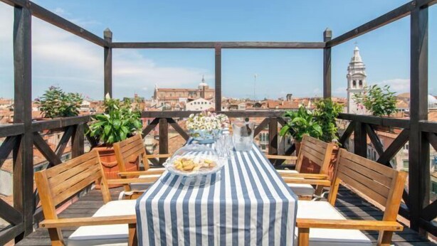 A rooftop terrace with a table set for two, surrounded by wooden chairs, potted plants, and a clear view of the cityscape. - Rooftop Terrace Penthouse & 360° Views Venice5th (Venice)