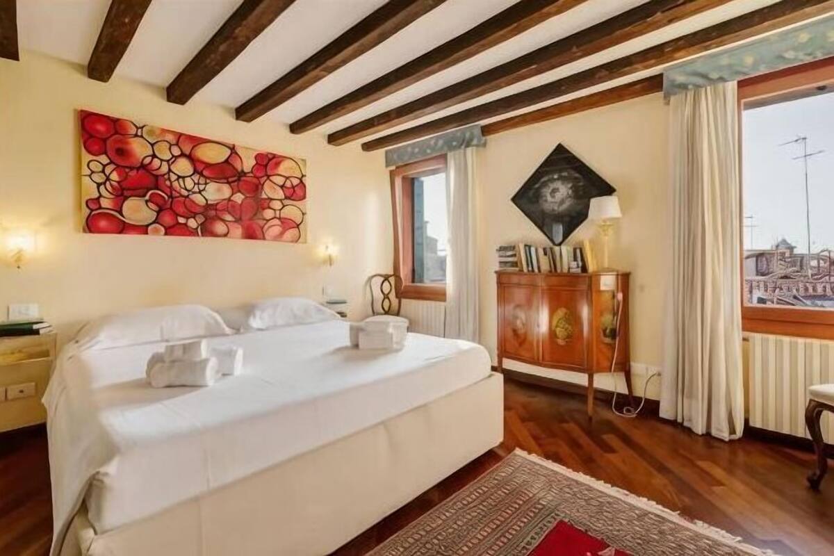 A hotel room with a bed, wooden beams, a painting, a cabinet, and a window with curtains.