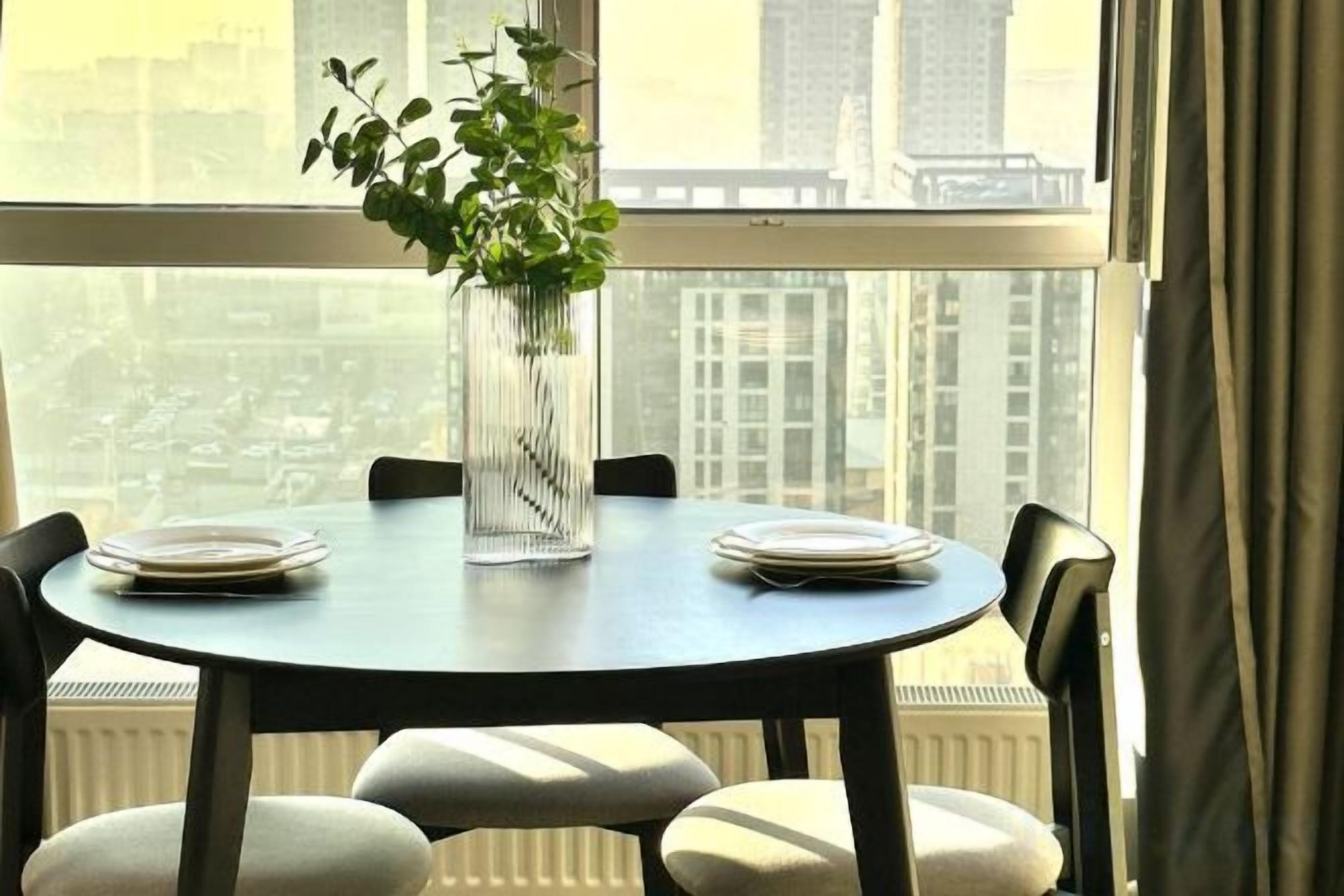A dining table with plates and a vase of greenery, set against a window with a city view.