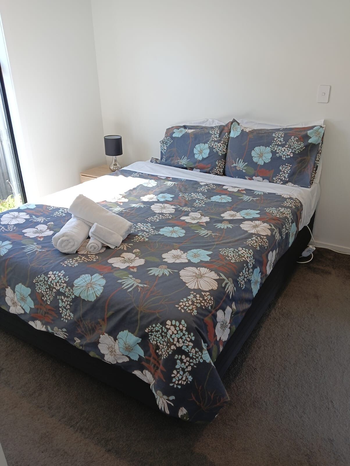 Una cama bien hecha con ropa de cama floral, una lámpara de noche y un enchufe en la pared.