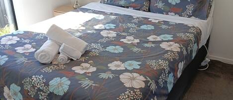Una cama bien hecha con ropa de cama floral, una lámpara de noche y un enchufe en la pared.