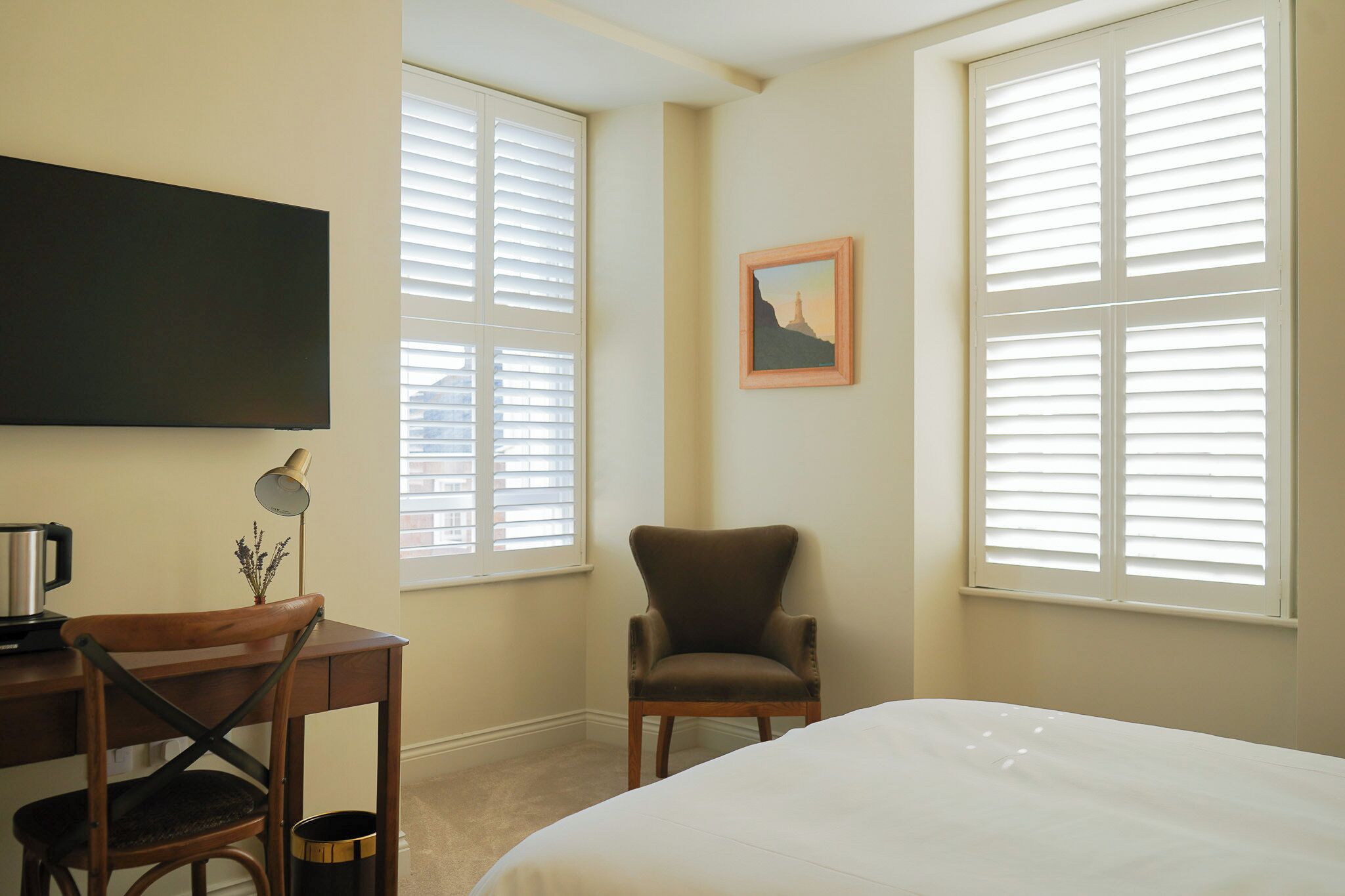 Une chambre d’hôtel avec un lit, un bureau, une chaise et deux fenêtres équipées de stores.