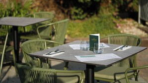 Un espace extérieur aménagé pour les repas, avec des chaises et des tables en métal vert, un menu et un verre d’eau posé sur la table.