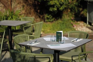 Un espace extérieur aménagé pour les repas, avec des chaises et des tables en métal vert, un menu et un verre d’eau posé sur la table.