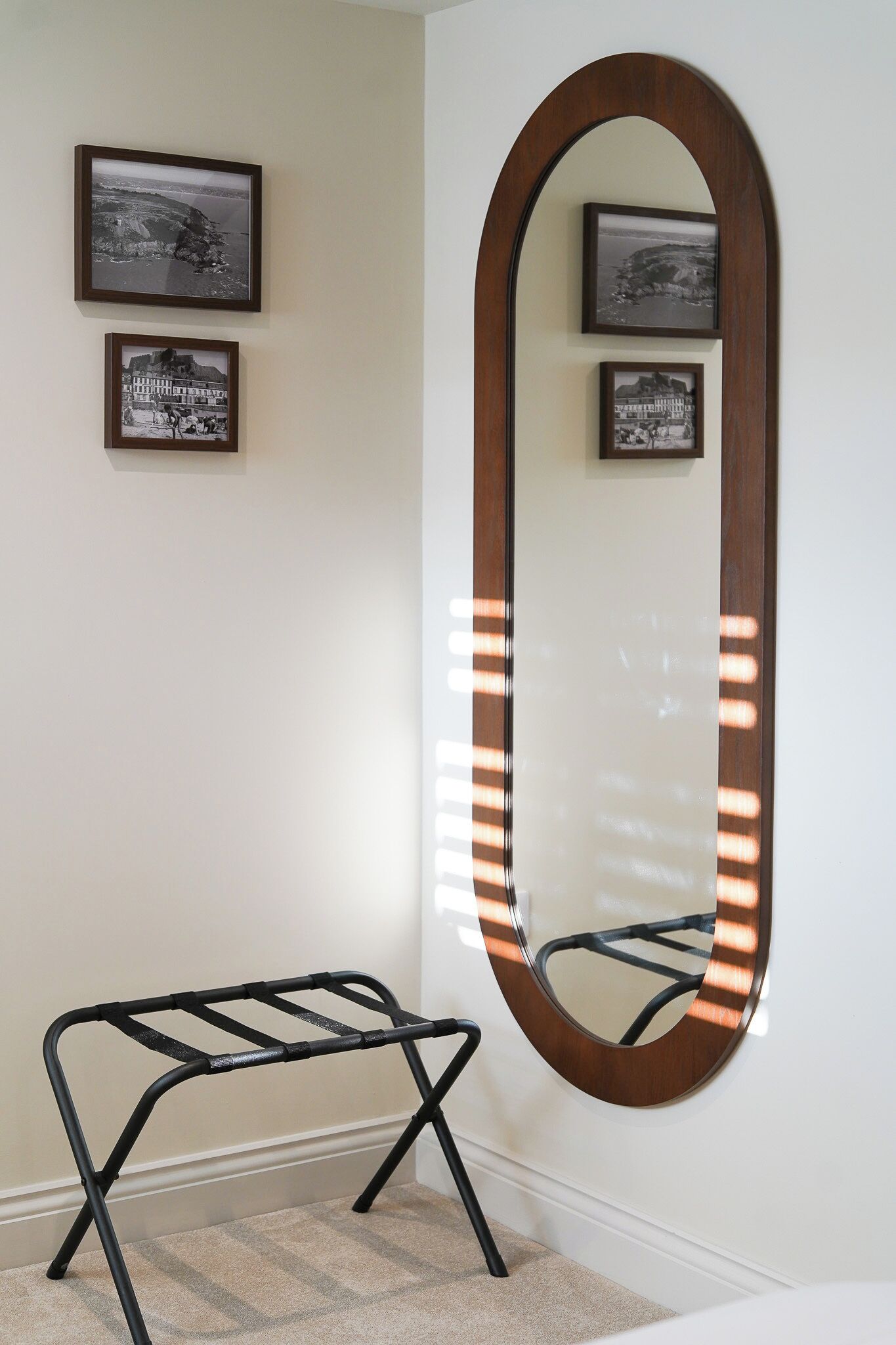 A room with a large oval mirror with lights, a black metal bench, and three framed pictures on the wall.